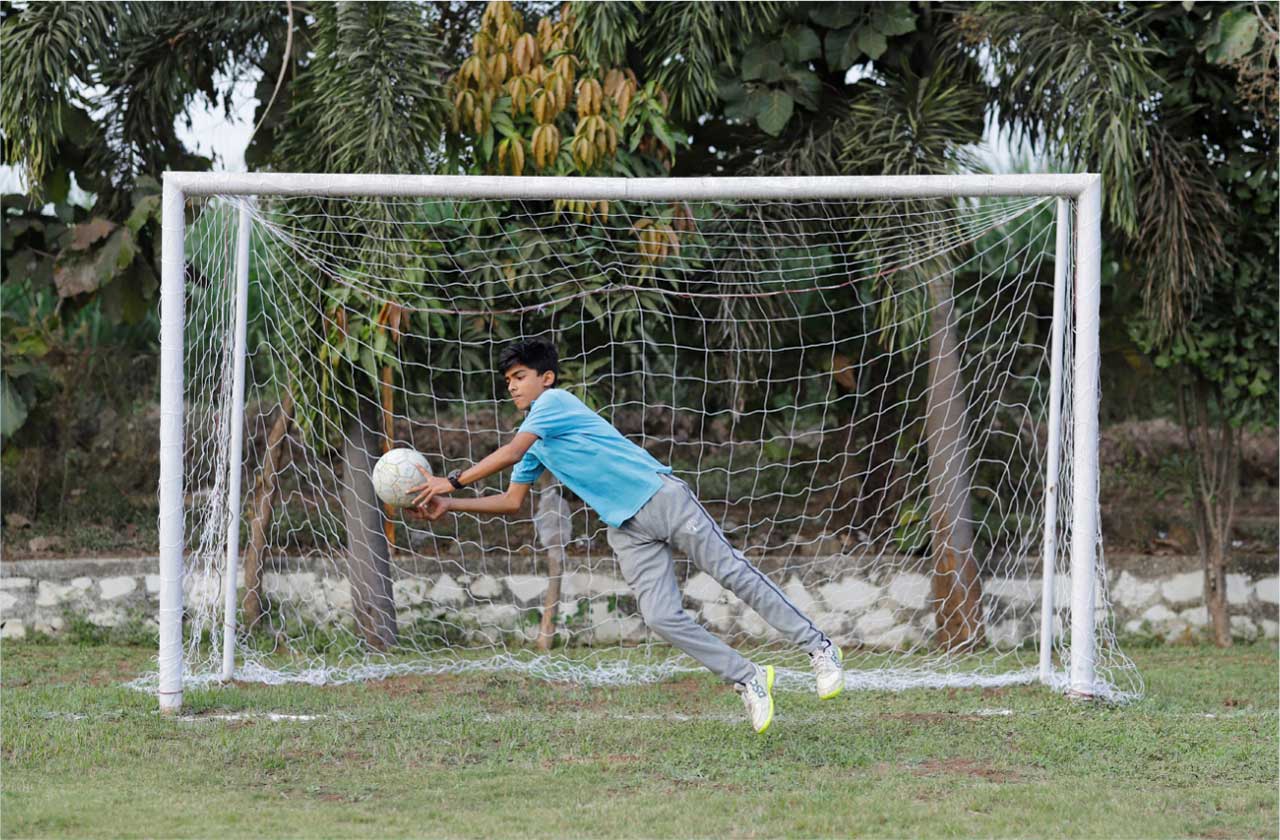 Students playing on a school playground (football/cricket) with a vibrant campus in the background.